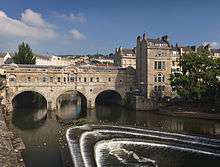 A three arch stone bridge with buildings on it, over water. Below the bridge is a three step weir and pleasure boat.