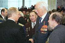 A man in the center, facing the left, is wearing medals on a jacket. He is shaking hands with another man, watched by three others.