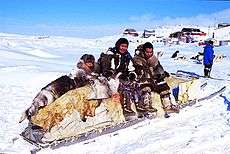 Color photograph of three Inuit persons on a traditional qamutik (sled), in a snow-covered landscape in Cape Dorset.