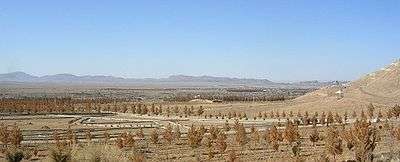 Sparse brown vegetation in dusty soil fills the foreground, fading to distant mountains along the horizon. A barely discernible scattering of buildings is in the middle-distance.