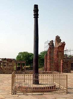 An pillar, slightly fluted, with some ornamentation at its top. It is black, slightly weathered to a dark brown near the base. It is around 7 meters (23 feet) tall. It stands upon a raised circular base of stone, and is surrounded by a short, square fence.