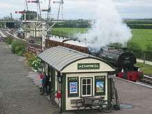 Steam locomotive at a curving station platform. On the platform is a small building with a curved roof.