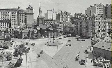 Aerial black and white photo of the junction of Macquarie and King Streets when traffic could still run from one into the other. Buses and other vehicles are on the roads and the now demolished building at the corner can be seen