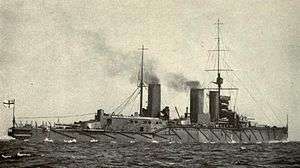 A dark-gray painted warship with two masts, three funnels, and four gun turrets visible at sea