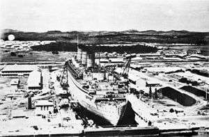A massive ocean liner, viewed from aft and above, sits in a dry dock.