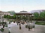A gazebo-structure with drinking fountains at the base, in a tree-lined square.