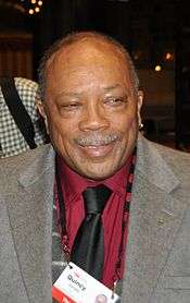 An elderly African-American man. The male is wearing a grey jacket open and a wine color shirt with a black tie.