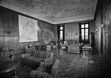 A black and white image of a large room, with two large windows on the back wall, with two more walls coming away from this wall at right angles. There are a number of large sofas spaced around the room, as well as single chairs, and a large desk surrounded by chairs. On the walls that do not have windows, one has a large map of northern Europe, whilst the other wall has a large doorway leading out of the room.