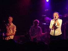 Linda Hopper singing with R.E.M.; singer Michael Stipe and bassist Mike Mills are visible to her left.