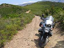 A BMW R1150GS parked on a gravel trail with scrub-covered mountains in the background