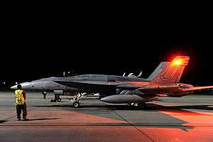 Side view of military combat jet with external fuel tanks and wingtip missiles, parked on airfield at night with ground crewman in foreground