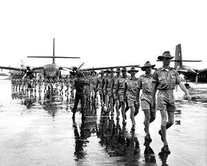 Uniformed personnel marching on airfield with two twin-engined transport aircraft in background