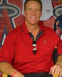 A man in a red shirt smiles with a pair of sunglasses folded in the front of his shirt.