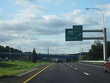 Ground-level view of a freeway; a large, green sign marked with "Exit 7" is posted directly overhead. Two large bridges are visible in the distance.