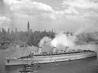 Black and white photograph showing the portside of the RMS Queen Mary entering New York Harbor on 20 June 1945 being accompanied by two small tug boats