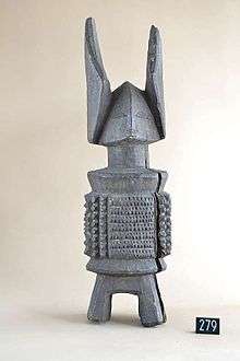 An image of a carved deity named Ikenga, the grey wooden piece has legs, a stylised but simple body, a trinagular head and shallow facial features and two horns around 1/3 its size