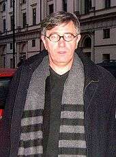 A middle-aged man with greying hair, wearing black-rimmed oval spectacles and a dark grey jacket and light grey and black striped scarf, photographed in the street