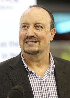 Rafael Benitez is seen with a beard while wearing a coat over a buttoned-up shirt.