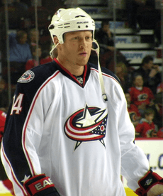 A Caucasian ice hockey player standing relaxed on the ice. He wears a white jersey with blue trim and a white helmet. He is looking forwards and holds his hockey stick with both hands across his torso.