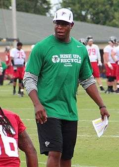 Candid photography of Morris on a football field wearing a green shirt bearing the slogan "Rise up & recycle", black shorts with an Atlanta Falcons logo and a white Falcons baseball cap holding a piece of paper in his left hand