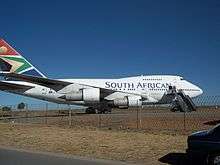 A Boeing 747SP, a shortened Boeing 747-100, is parked at an fenced-off airport, facing right. The aircraft's engines feature prominently, as a mobile stairway is placed next to one of its doors under the "N" in South African.