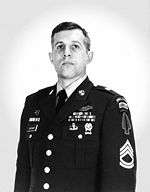 Head and torso of a white man standing erect and looking upwards, wearing a military jacket with an assortment of ribbon bars and badges on the left breast and chevrons and patches on the upper sleeve.