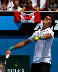 Raonic looks up in the air, his racquet pointing down in his left hand and a ball cradled in his right hand. In the background, a spectator holds a Canadian flag.