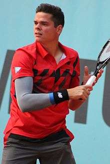 Closeup of Raonic, wearing a red short-sleeved shirt, a grey sleeve on his right arm, and black wristbands. He is holding his racquet up to the right in both hands, looking left.