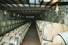 Colour photo showing an aging cellar for Sauternes (sweet white wine).  The barrels are aligned in three rows against the walls and they are stacked on two levels with a row in the middle one barrel high.  The light coloured barrels are newer barrels.  The floor is tiled, the walls are stone, covered in grey mould, and the ceiling is white supported by dark coloured beams.  The wall lighting is reminiscent of medieval torches.
