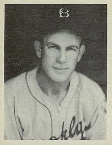 A man looks into the camera, smiling, while wearing a white baseball jersey and dark cap with a "B" on the center.