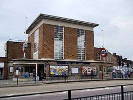 A brown-bricked building with a rectangular, dark blue sign reading "RAYNERS LANE STATION" in white letters all under a dark blue sky