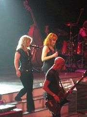 Image of two blond women walking down a step of stairs on the stage. An image of guitarist is seen standing next to them. Behind them, a set of drum and a cello are seen.