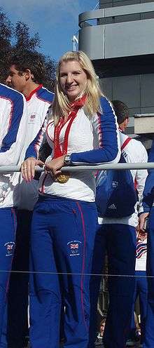 Young woman in blue tracksuit pants, white top with blue long sleeves, wearing a gold medal around her neck with red ribbon. She has long blonde hair, large nose and is leaning on a rail, and is surrounded by similarly dressed people.