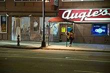 A woman in a brown fur coat stands smoking on the sidewalk of a city street, her hand resting on a pole supporting a red awning outside the entrance of a bar with a large red sign featuring the white words "Augie's".