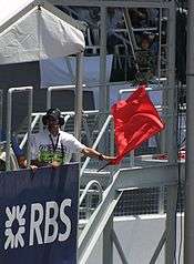 A man in a white overall, standing behind a blue placard with the letters RBS in white. He is waving a red flag.
