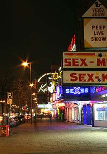 The Reeperbahn nighclub as seen from the side walk with its entrance lighted. Theatre marques say "Sex" and "Peep live shows".