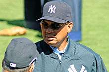 An African American male in his sixties wearing a cap and jacket, both with a logo of an overlapping "N" and two overlapping "Y"s, and sunglasses, stands on a baseball field.