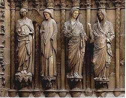 Four carved figures are set between columns and are mounted against the wide angled jamb of a medieval door. The figures are paired to illustrate two stories. To the left, a smiling angel approaches Mary who raises her hand. in the right groupt, Mary approaches her elderly cousin Elizabeth who turns her eyes to heaven. The style of the two groups is entirely different, the first being simpler and more elegant, the second having very elaborate drapery and realistic detail.
