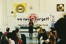 A seated crowd facing a standing woman. Behind her is a table with flowers. Above the table is a large banner with the text, "We never forget!" along with the IWW name and globe logo. A variety of United Auto Workers logos are visible on the wall in the background.