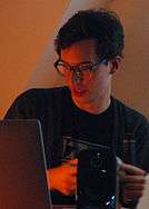 Bédard is holding an obfuscated controller while looking down and left at a computer screen. He has short, dark hair, medium-sized glasses, and wears a black t-shirt. The image has a warm palette and is grainy from the low light conditions.