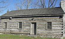A log structure with a door in the center, three windows, and a brick chimney on the right