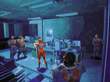 A young woman in a red and white racing suit is shooting at a pair of undead police officers, while two more enemies sneak up on her from behind. The office of the police station they are fighting in has sheets of paper scattered on the floor, and the whole scene features a bluish tint.