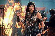 Xena, clad in leather armour accented and decorated in bronze detailing, standing in the middle of a battlefield, flames behind her as she holds her chakram in readiness for battle.