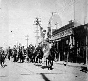Black-and-white photograph of a group of men on horseback on a city street. Lead horsemen is carrying a flag.