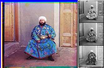 A large color photograph abutting (to its right) a column of three stacked black-and-white versions of the same picture.  Each of the three smaller black-and-white photos are slightly different, due to the effect of the color filter used.  Each of the four photographs differ only in color and depict a turbaned and bearded man, sitting in the corner an empty room, with an open door to his right and a closed door to his left.  The man is wearing an ornate full-length blue robe trimmed with a checkered red-and-black ribbon.  The blue fabric is festooned with depictions of stems of white, purple, and blue flowers.  He wears an ornate gold belt, and in his left hand he holds a gold sword and scabbard. Under his right shoulder strap is a white aiguillette; attached to his robe across his upper chest are four multi-pointed badges of various shapes, perhaps military or royal decorations.