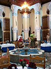 A large, open Moroccan riad that is filled with dining tables and chairs. In the center of the room is a fountain shaped like a flower with eight petals. A chandelier hangs from the ceiling, which is supported by three columns at the back. A group of people eat a meal at the table closest to the columns.