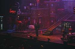 A man standing in front of the audience while in the middle of a concert, surrounded by the apartment-styled scenography and the rest of the musicians.
