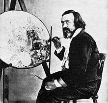 Photograph of a bearded, middle-aged man. He is seated and holding a paint brush. In front of him is an unfinished, circular painting on an artist's easel.