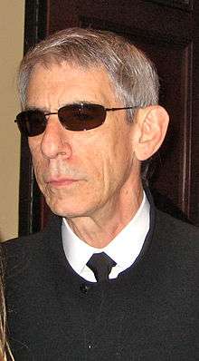 An unsmiling gray-haired man wearing sunglasses and a black shirt with a white collar looks off-screen.