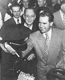 A smiling young man shakes hands; a Nixon bumper sticker is seen.